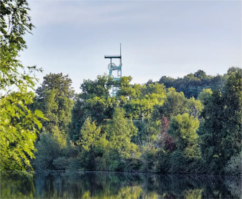 Ehemaliger Förderturm eines Steinkohlebergwerks, umgeben von dichtem Grün, spiegelt sich in einem See im Vordergrund.
