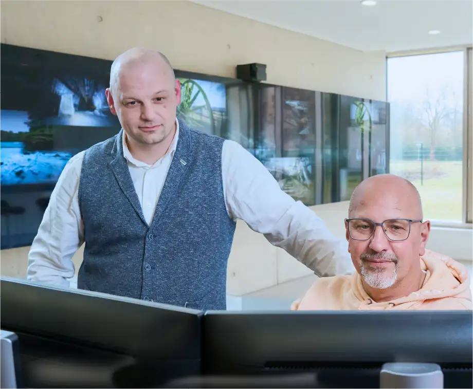 Zwei Männer schauen gemeinsam auf Bildschirme in einem Kontrollraum, im Hintergrund großformatige Monitore mit Bergbau- und Infrastrukturaufnahmen.