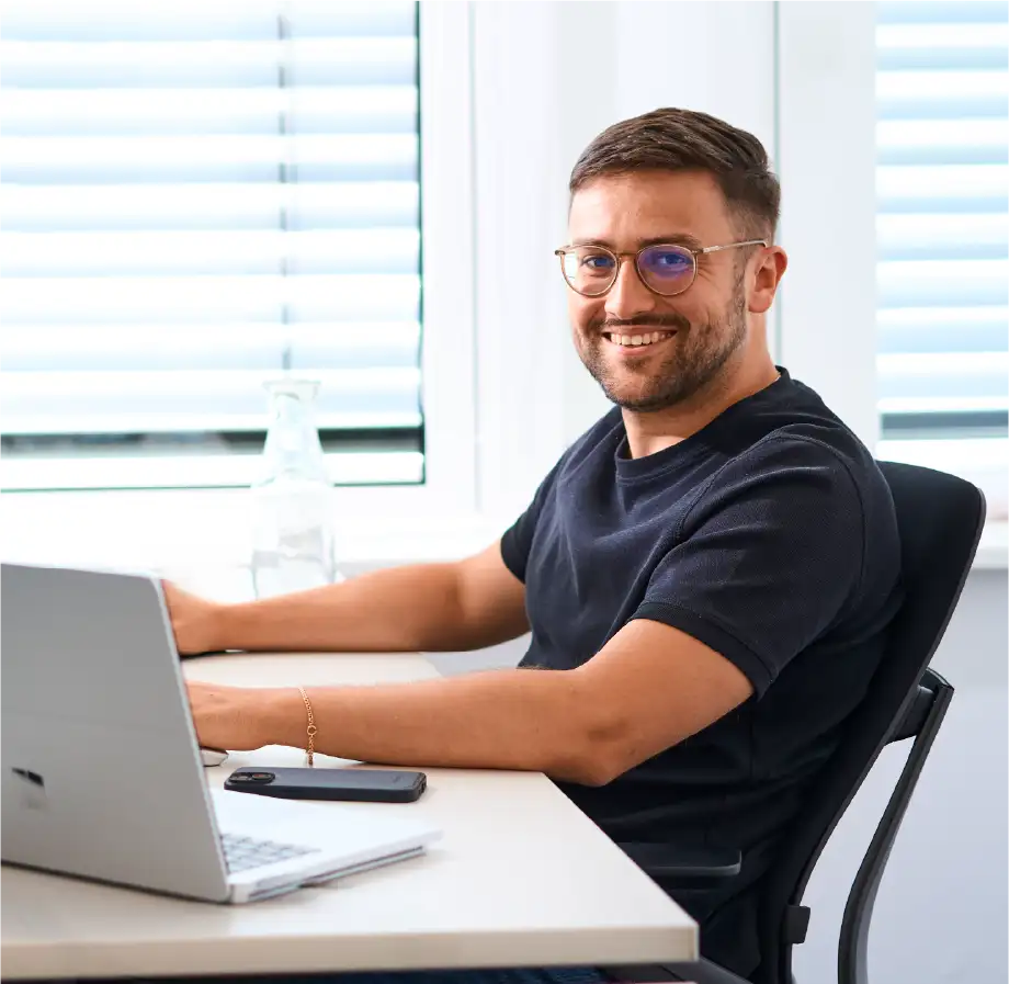 Mann mit Brille sitzt lächelnd am Laptop in einem hellen Büro mit Fensterjalousien im Hintergrund.